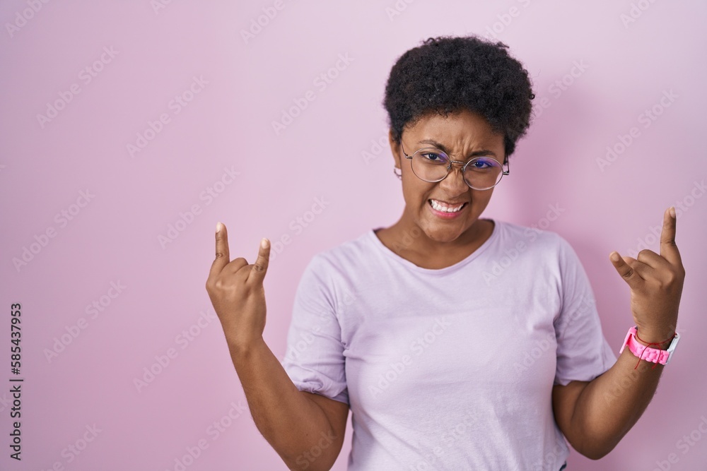 Young african american woman standing over pink background shouting with crazy expression doing rock symbol with hands up. music star. heavy concept. Young african american woman standing over pink background shouting with crazy expression doing rock symbol with hands up. music star. heavy concept.