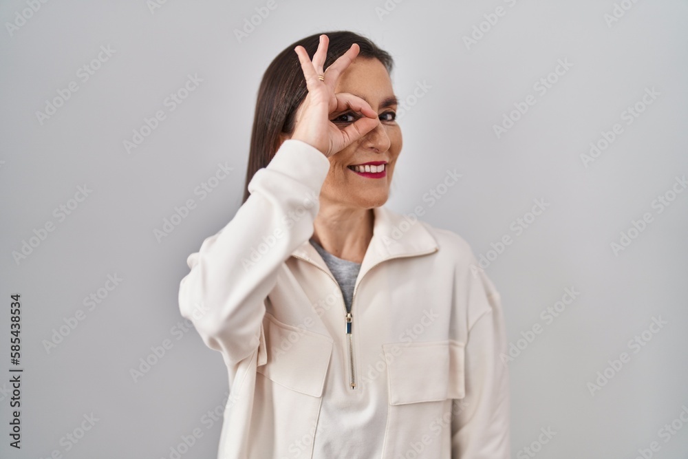 Middle age hispanic woman standing over isolated background doing ok gesture with hand smiling, eye looking through fingers with happy face.