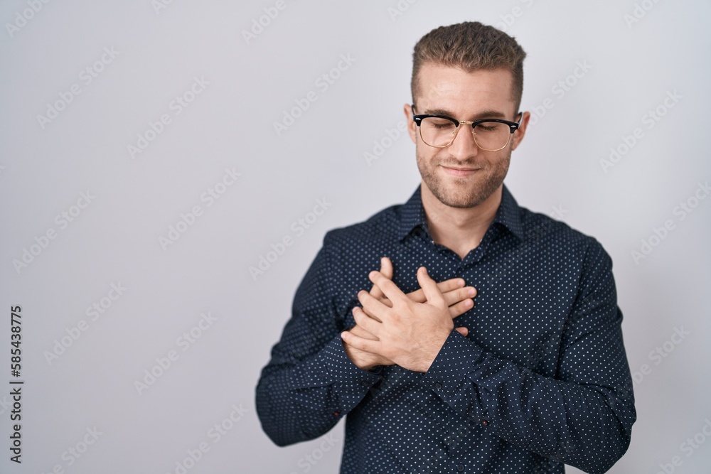 Young caucasian man standing over isolated background smiling with hands on chest with closed eyes and grateful gesture on face. health concept. Young caucasian man standing over isolated background smiling with hands on chest with closed eyes and grateful gesture on face. health concept.