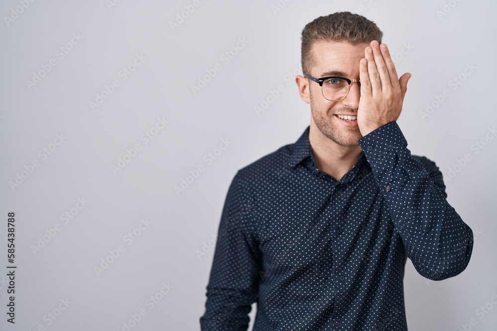 Young caucasian man standing over isolated background covering one eye with hand, confident smile on face and surprise emotion. Young caucasian man standing over isolated background covering one eye with hand, confident smile on face and surprise emotion.