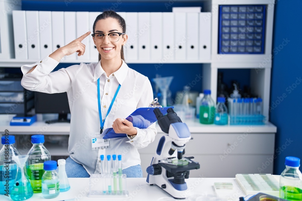 Young brunette woman working at scientist laboratory smiling pointing to head with one finger, great idea or thought, good memory Young brunette woman working at scientist laboratory smiling pointing to head with one finger, great idea or thought, good memory