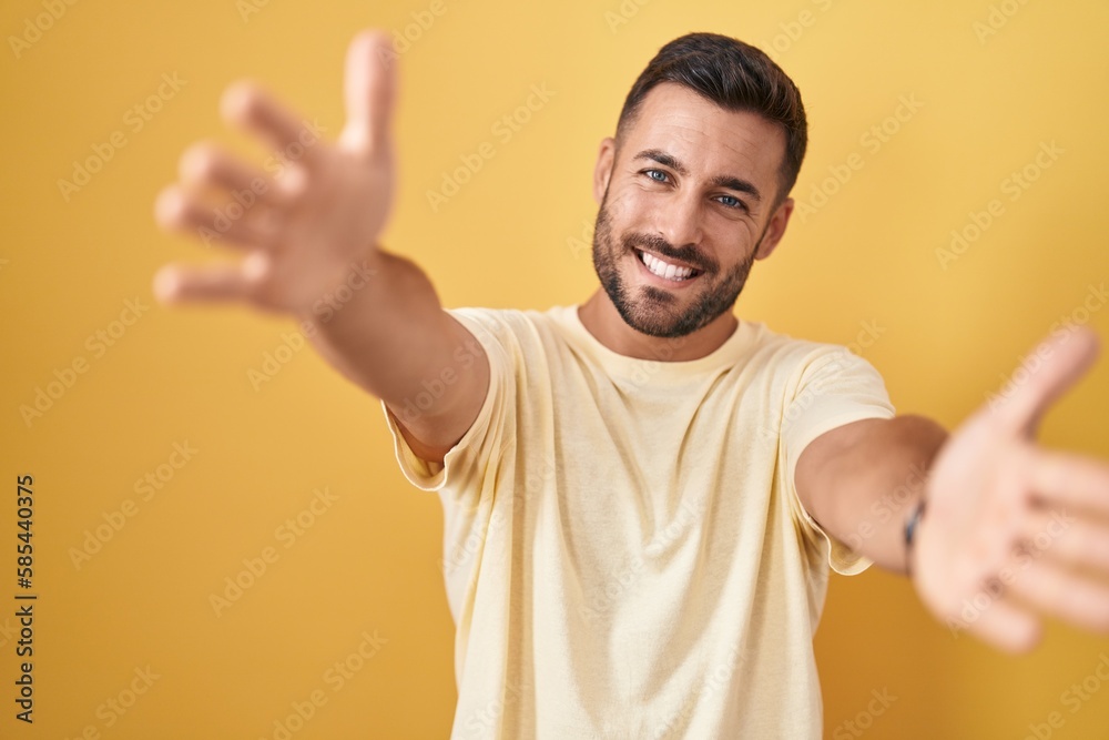 Handsome hispanic man standing over yellow background looking at the camera smiling with open arms for hug. cheerful expression embracing happiness. Handsome hispanic man standing over yellow background looking at the camera smiling with open arms for hug. cheerful expression embracing happiness.