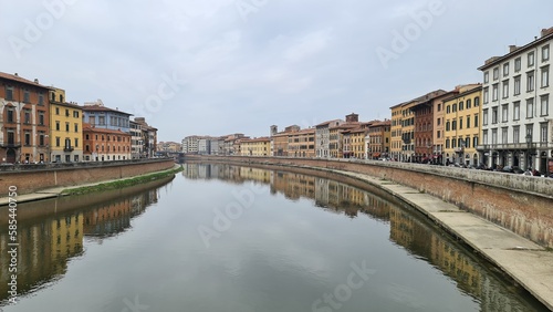 Pisa, Italy - February 25, 2023: View of the medieval town of Pisa from bridge "Ponte di Mezzo" on river Arno in winter days, with grey sky in the background. Reflection of the buildings on the water.