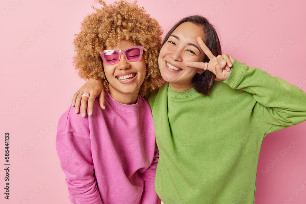 © wayhome.studio - Horizontal shot of positive mixed race young women friends smile and embrace make peace gesture have happy expressions dressed casually isolated over pink background. People and emotions concept © wayhome.studio - Horizontal shot of positive mixed race young women friends smile and embrace make peace gesture have happy expressions dressed casually isolated over pink background. People and emotions concept