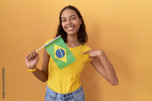 Young hispanic woman holding brazil flag looking confident with smile on face  pointing oneself with fingers proud and happy.