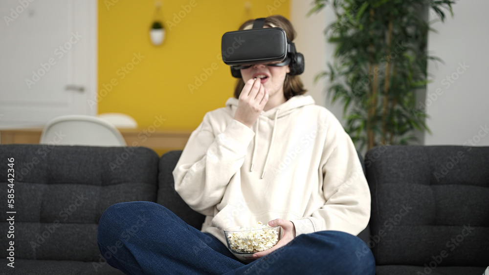 Young blonde woman watching movie using virtual reality glasses eating popcorn at home