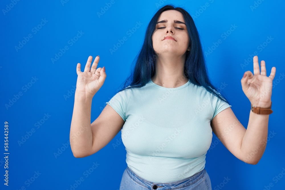 Young modern girl with blue hair standing over blue background relaxed and smiling with eyes closed doing meditation gesture with fingers. yoga concept. Young modern girl with blue hair standing over blue background relaxed and smiling with eyes closed doing meditation gesture with fingers. yoga concept.