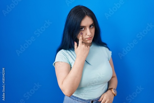 Young modern girl with blue hair standing over blue background pointing to the eye watching you gesture, suspicious expression © Krakenimages.com
