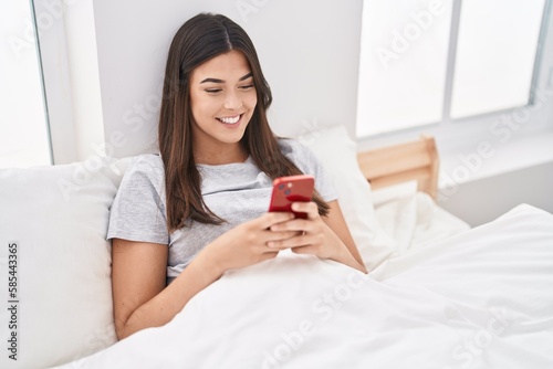 Young beautiful hispanic woman using smartphone sitting on bed at bedroom