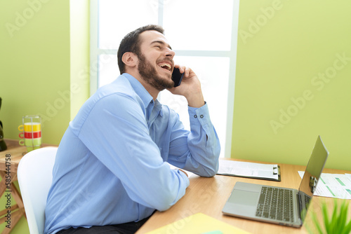 Young hispanic man business worker using laptop talking on smartphone at office