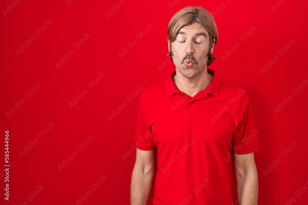 Fototapeta premium Caucasian man with mustache standing over red background making fish face with lips, crazy and comical gesture. funny expression.
