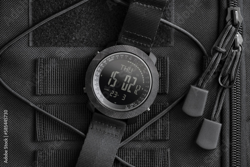 Tactical watch on backpack. Military field electronic watch in black steel case, having notched bezel, built-in compass, with nylon strap, lies on black tactical backpack, close-up.