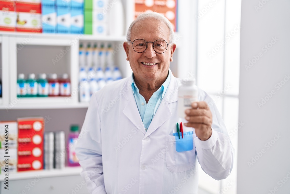 Senior grey-haired man pharmacist smiling confident holding pills bottle at pharmacy