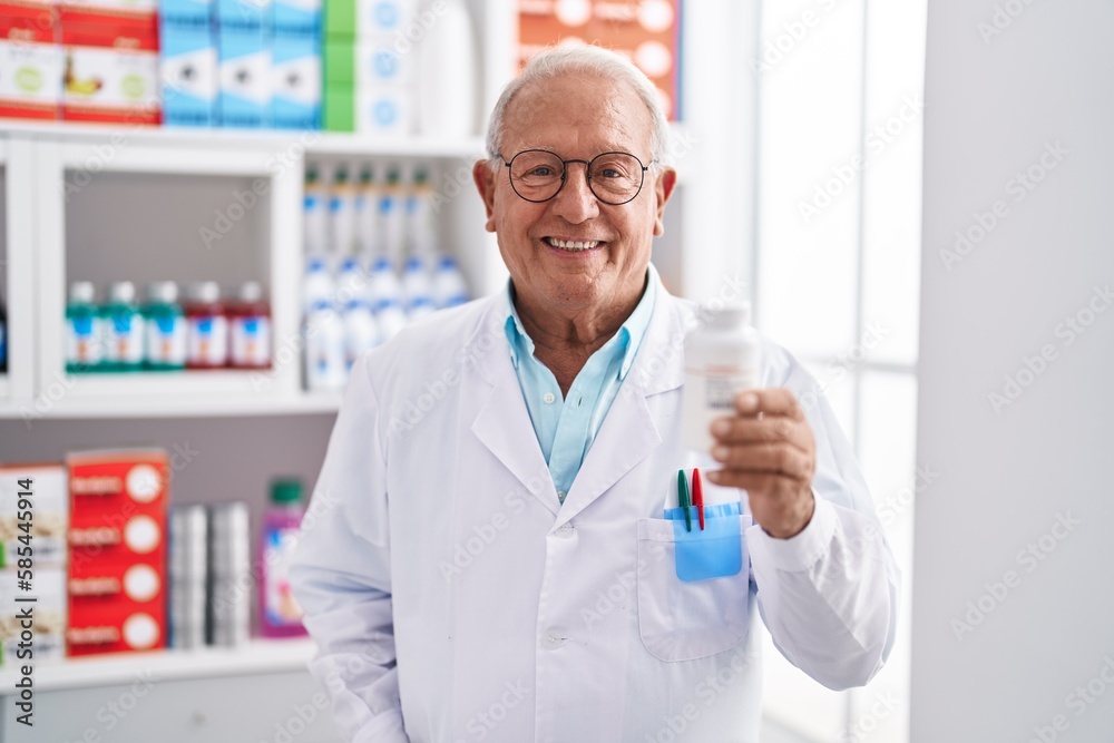 Senior grey-haired man pharmacist smiling confident holding pills bottle at pharmacy Senior grey-haired man pharmacist smiling confident holding pills bottle at pharmacy
