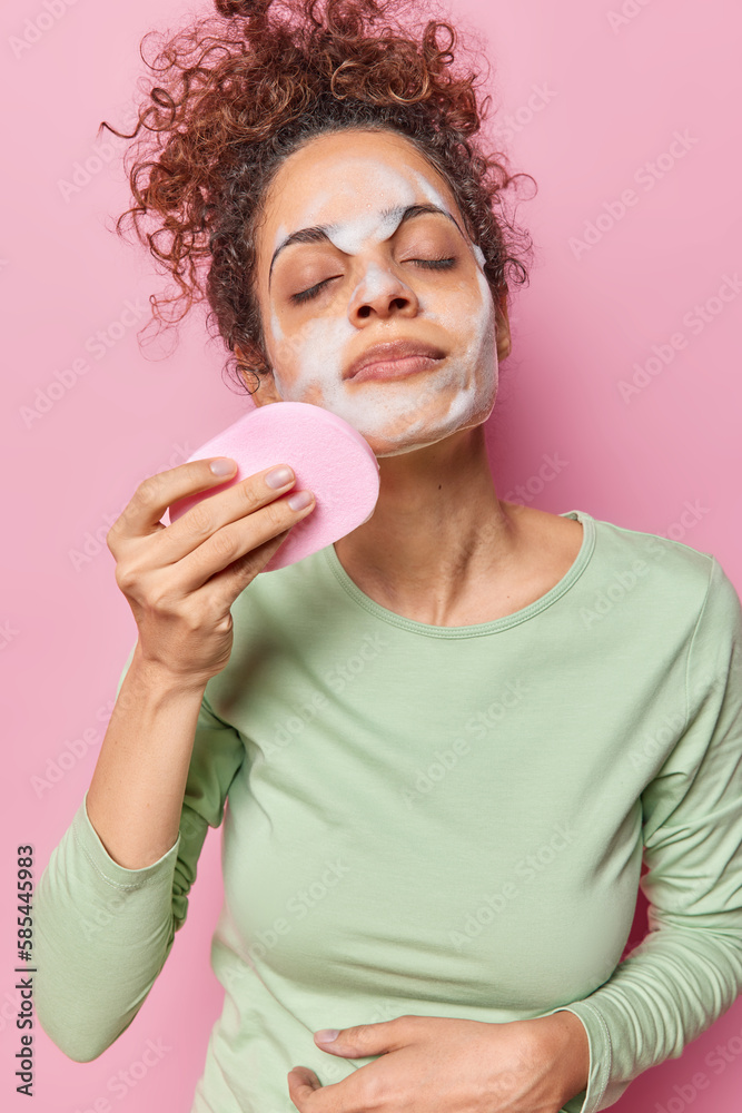 Photo of teenage girl with curly hair refreshes skin undergoes beauty procedures applies foam gel with sponge dressed in casual jumper enjoys purity satisfied with purity of skin. Studio shot Photo of teenage girl with curly hair refreshes skin undergoes beauty procedures applies foam gel with sponge dressed in casual jumper enjoys purity satisfied with purity of skin. Studio shot