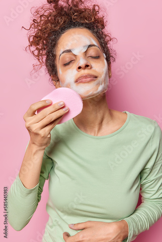 Photo of teenage girl with curly hair refreshes skin undergoes beauty procedures applies foam gel with sponge dressed in casual jumper enjoys purity satisfied with purity of skin. Studio shot