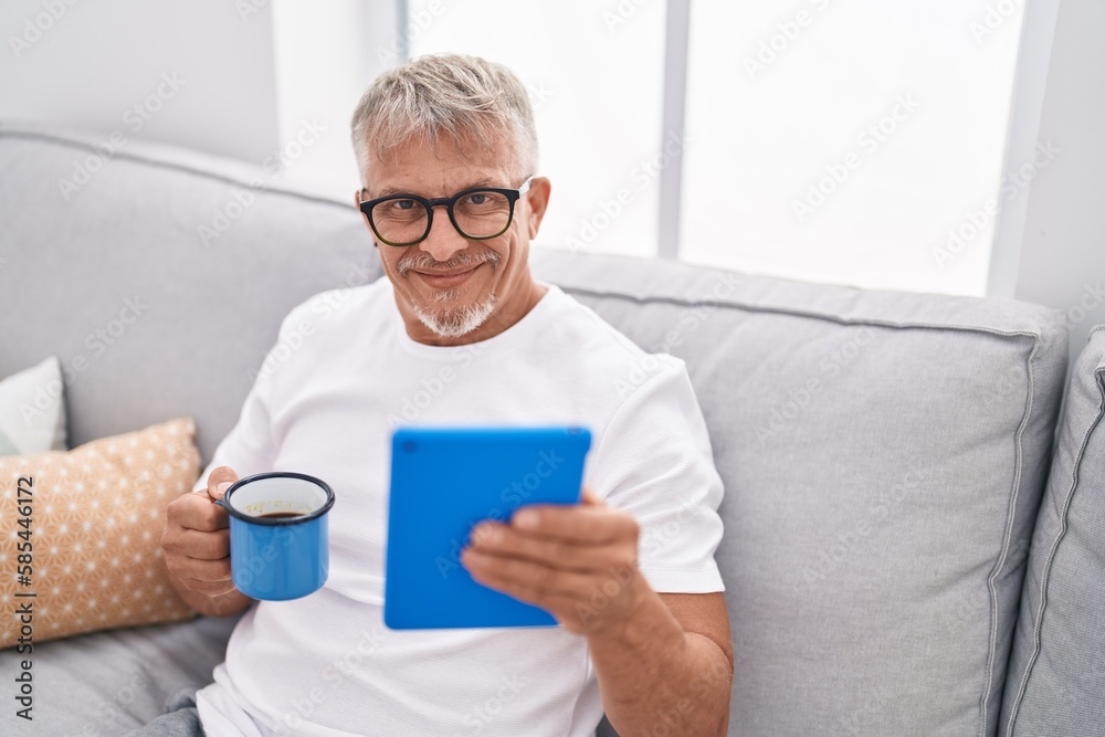 Middle age grey-haired man using touchpad drinking coffee at home Middle age grey-haired man using touchpad drinking coffee at home