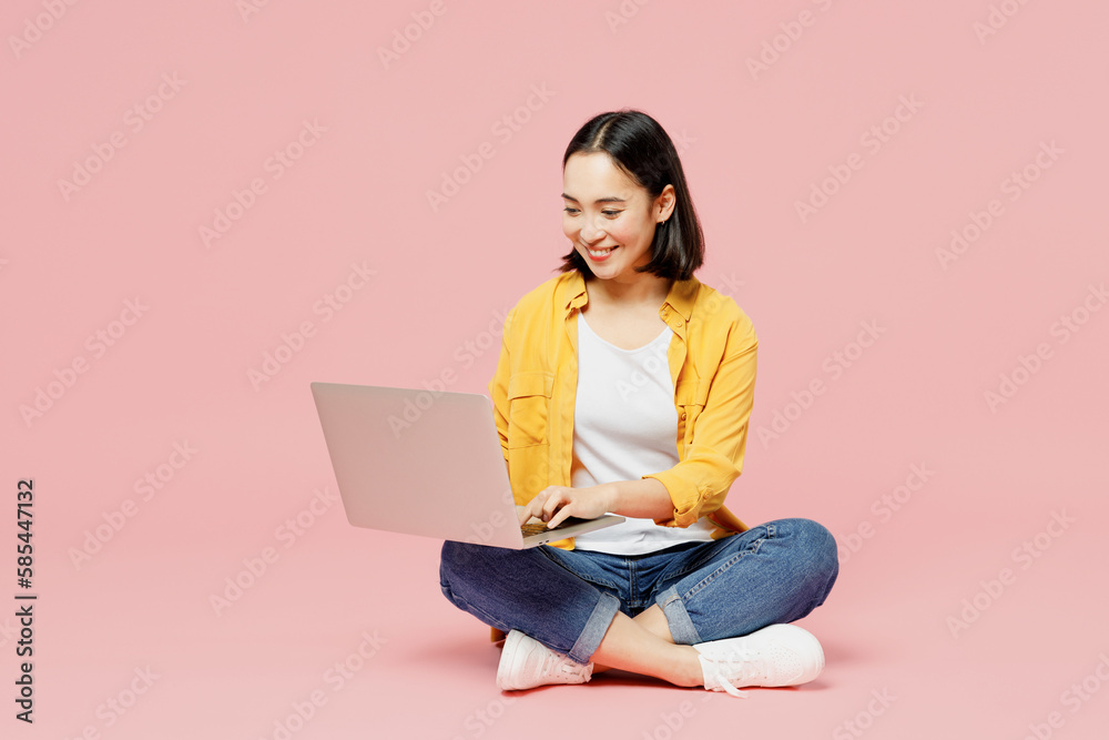 Full body young woman of Asian ethnicity wear yellow shirt white t-shirt sitting hold use work on laptop pc computer isolated on plain pastel light pink background studio portrait. Lifestyle concept. Full body young woman of Asian ethnicity wear yellow shirt white t-shirt sitting hold use work on laptop pc computer isolated on plain pastel light pink background studio portrait. Lifestyle concept.