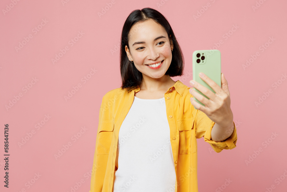 Young happy woman of Asian ethnicity wear yellow shirt white t-shirt doing selfie shot on mobile cell phone post photo on social network isolated on plain pastel light pink background studio portrait. Young happy woman of Asian ethnicity wear yellow shirt white t-shirt doing selfie shot on mobile cell phone post photo on social network isolated on plain pastel light pink background studio portrait.
