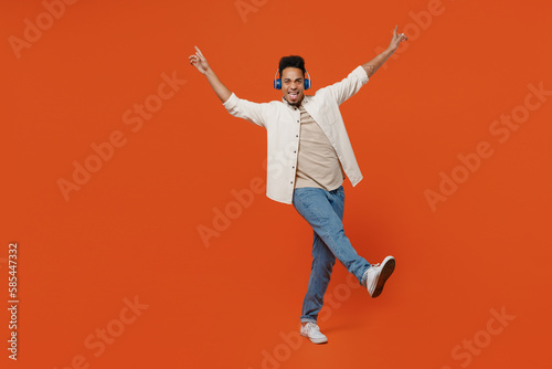 Full body young man of African American ethnicity wear light shirt casual clothes headphones listen music spread hands raise up leg isolated on orange red background studio portrait Lifestyle concept