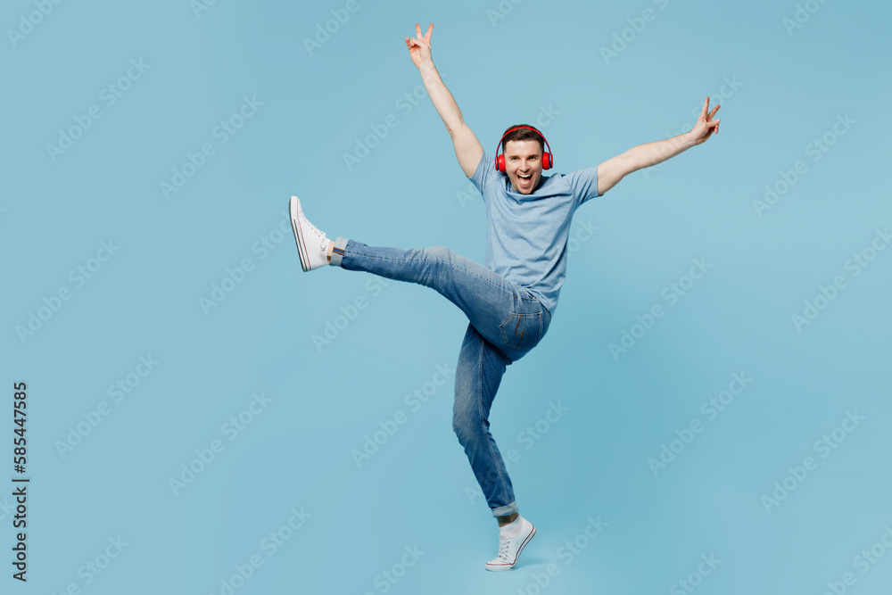 Full body young smiling man wear casual t-shirt headphones listen to music with outstretched hands raise up leg isolated on plain pastel light blue cyan background studio portrait. Lifestyle concept.