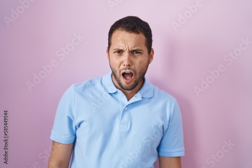 Hispanic man standing over pink background in shock face, looking skeptical and sarcastic, surprised with open mouth