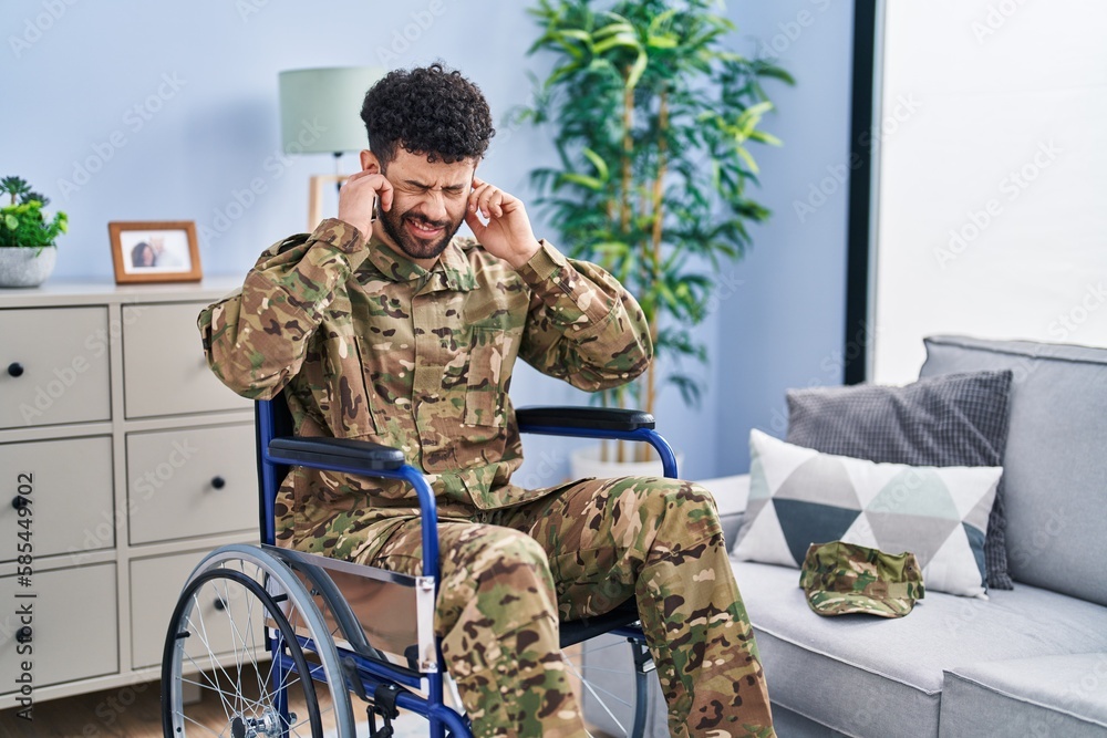 Fototapeta premium Arab man wearing camouflage army uniform sitting on wheelchair covering ears with fingers with annoyed expression for the noise of loud music. deaf concept.