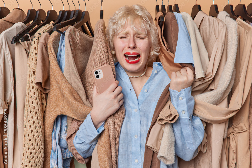 Upset crying woman feels frustrated stands near her wardrobe has nothing to wear and no money for buying new outfit holds smartphone poses around clothing on hangers. Unsuccessful shopping day