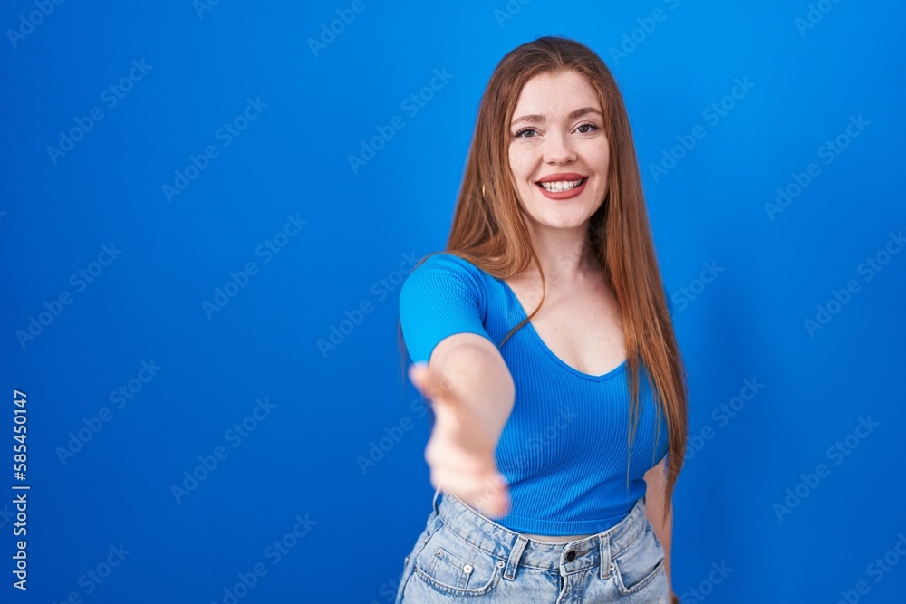 Redhead woman standing over blue background smiling friendly offering handshake as greeting and welcoming. successful business. Redhead woman standing over blue background smiling friendly offering handshake as greeting and welcoming. successful business.