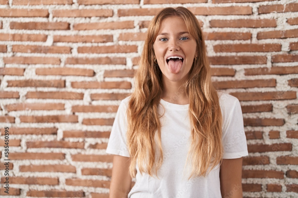 Young caucasian woman standing over bricks wall sticking tongue out happy with funny expression. emotion concept. Young caucasian woman standing over bricks wall sticking tongue out happy with funny expression. emotion concept.