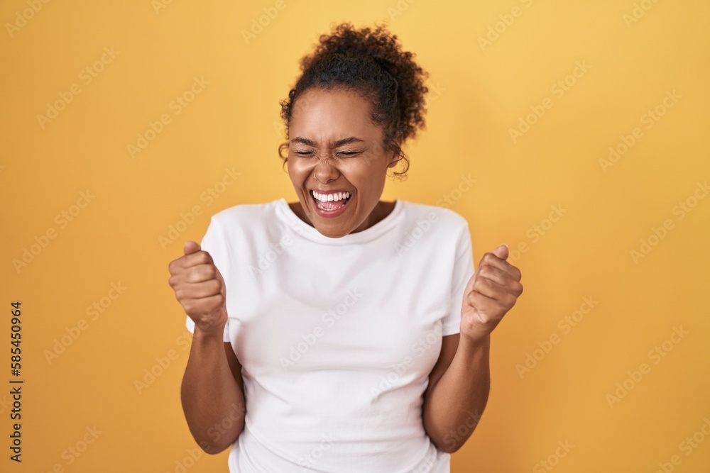 Young hispanic woman with curly hair standing over yellow background excited for success with arms raised and eyes closed celebrating victory smiling. winner concept. Young hispanic woman with curly hair standing over yellow background excited for success with arms raised and eyes closed celebrating victory smiling. winner concept.