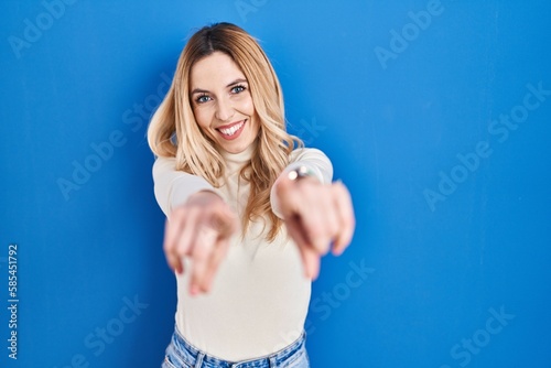 Young caucasian woman standing over blue background pointing to you and the camera with fingers, smiling positive and cheerful