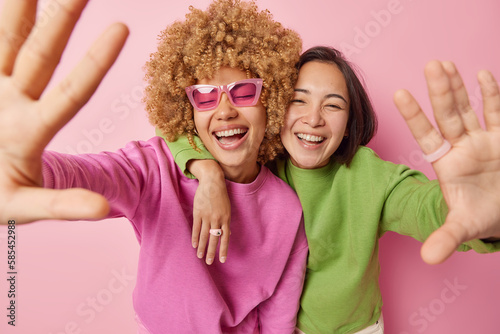 Positive overjoyed women keep arms outstreched laugh happily dressed in green and pink jumpers embrace and foolish around smile gladfully poses indoor. Friendship positive emotions and fun concept