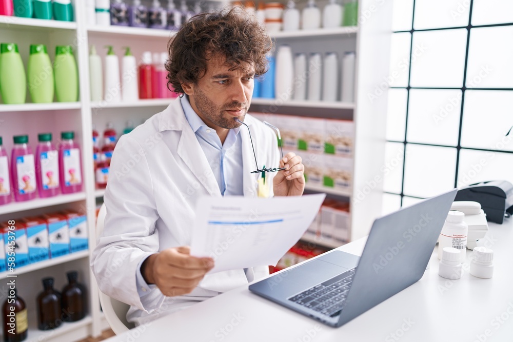 Young hispanic man pharmacist reading prescription using laptop at pharmacy