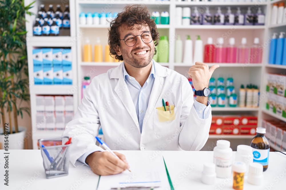 Hispanic young man working at pharmacy drugstore smiling with happy face looking and pointing to the side with thumb up.