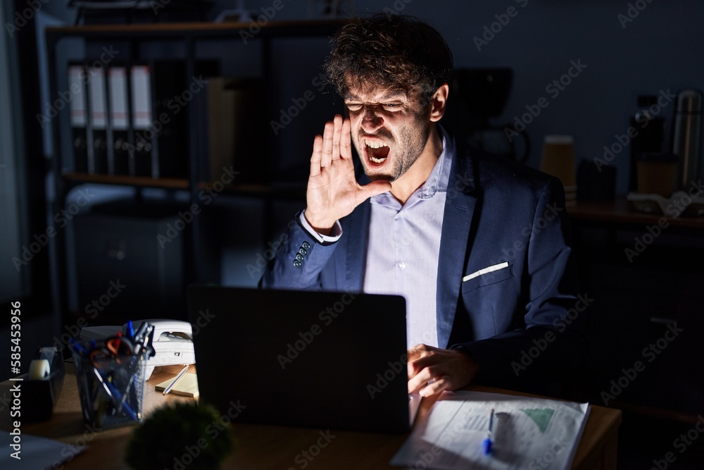 Hispanic young man working at the office at night shouting and screaming loud to side with hand on mouth. communication concept. Hispanic young man working at the office at night shouting and screaming loud to side with hand on mouth. communication concept.