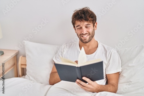 Young man reading book sitting on bed at bedroom