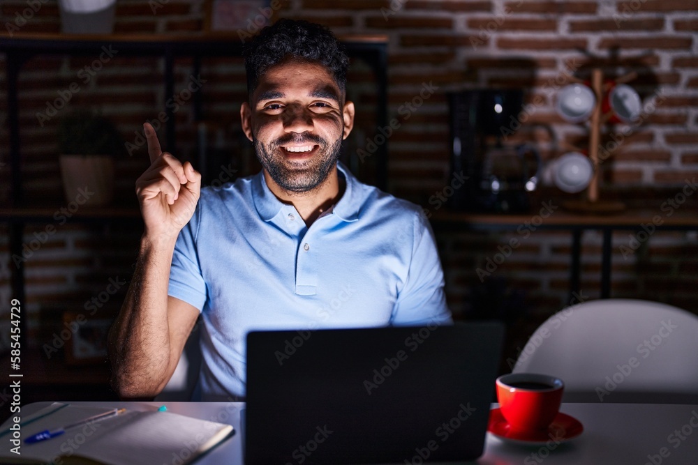 Hispanic man with beard using laptop at night with a big smile on face, pointing with hand and finger to the side looking at the camera. Hispanic man with beard using laptop at night with a big smile on face, pointing with hand and finger to the side looking at the camera.