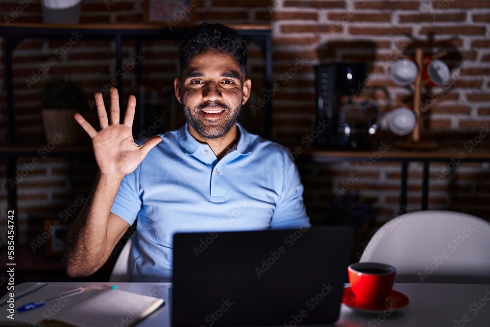 Hispanic man with beard using laptop at night showing and pointing up with fingers number five while smiling confident and happy. Hispanic man with beard using laptop at night showing and pointing up with fingers number five while smiling confident and happy.