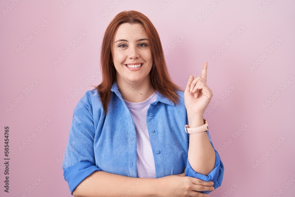 Young hispanic woman with red hair standing over pink background with a big smile on face, pointing with hand finger to the side looking at the camera. Young hispanic woman with red hair standing over pink background with a big smile on face, pointing with hand finger to the side looking at the camera.