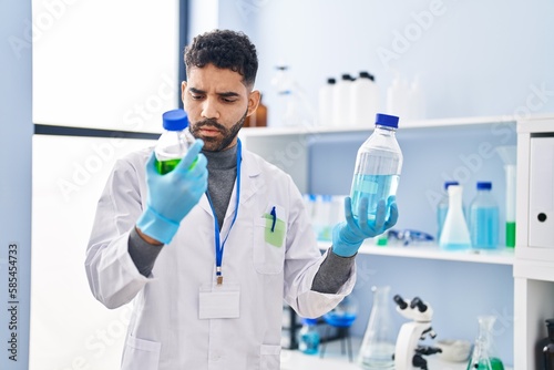 Young hispanic man wearing scientist uniform holding bottles at laboratory