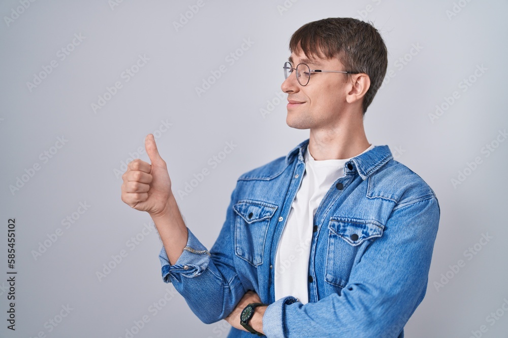 Caucasian blond man standing wearing glasses looking proud, smiling doing thumbs up gesture to the side Caucasian blond man standing wearing glasses looking proud, smiling doing thumbs up gesture to the side