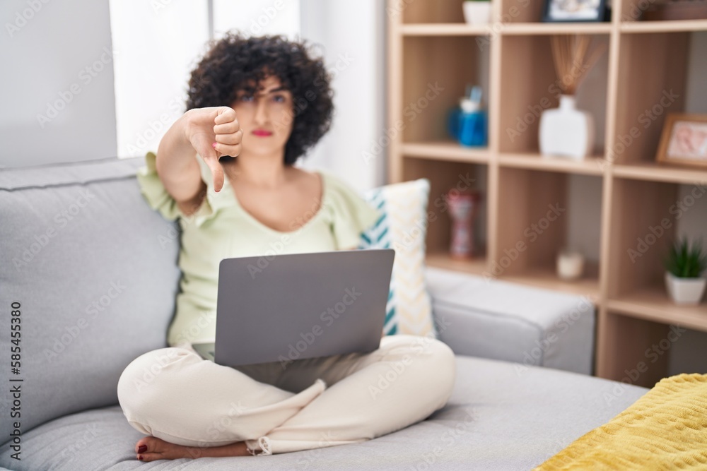 Young brunette woman with curly hair using laptop sitting on the sofa at home looking unhappy and angry showing rejection and negative with thumbs down gesture. bad expression.