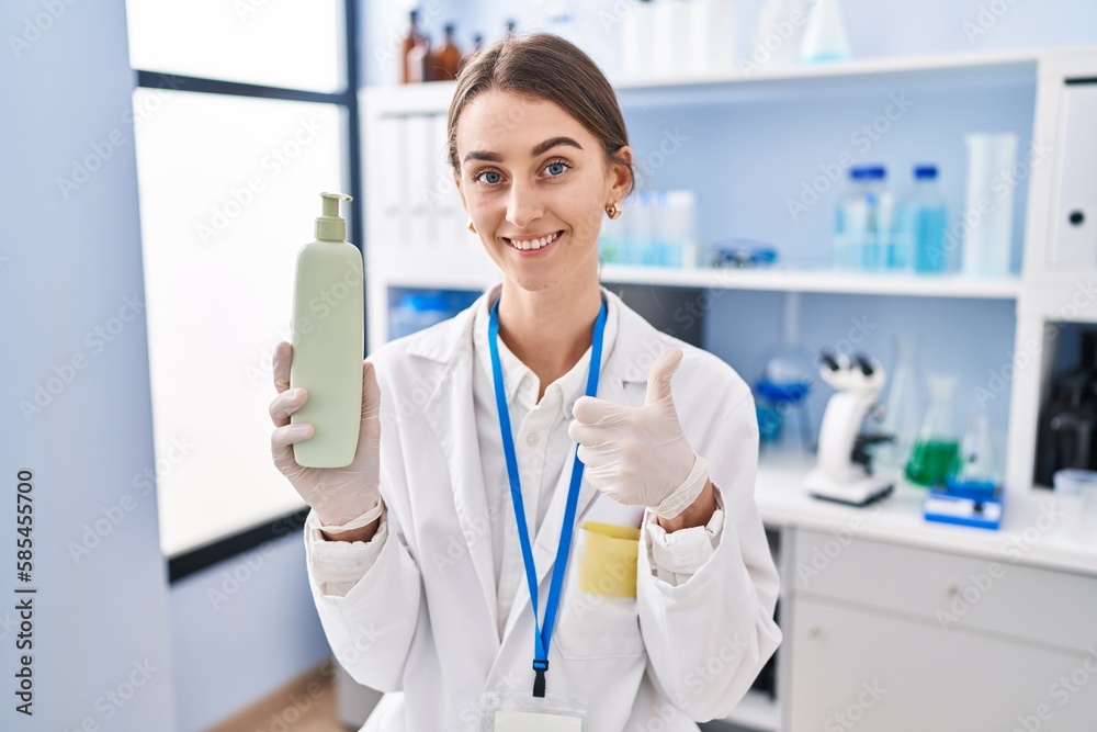 Young caucasian woman working at scientist laboratory holding body lotion smiling happy and positive, thumb up doing excellent and approval sign Young caucasian woman working at scientist laboratory holding body lotion smiling happy and positive, thumb up doing excellent and approval sign