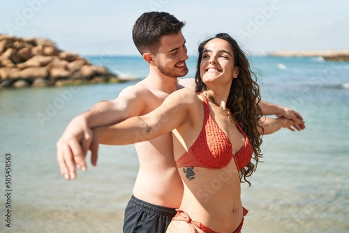 Young hispanic couple tourists wearing swimsuit standing with arms open at seaside