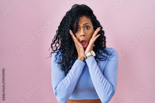 Middle age hispanic woman standing over pink background afraid and shocked, surprise and amazed expression with hands on face
