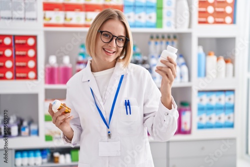 Young blonde woman pharmacist smiling confident holding pills bottles at pharmacy