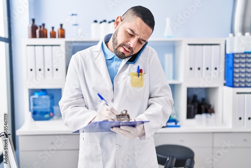Young latin man scientist talking on smartphone writing document at laboratory