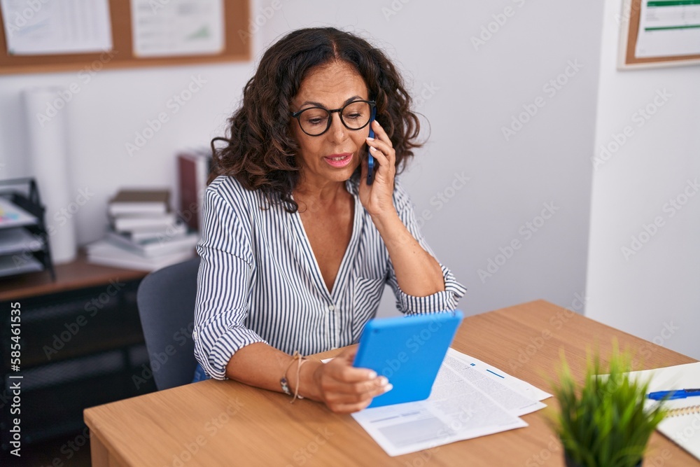 Middle age woman business worker talking on smartphone using touchpad at office Middle age woman business worker talking on smartphone using touchpad at office