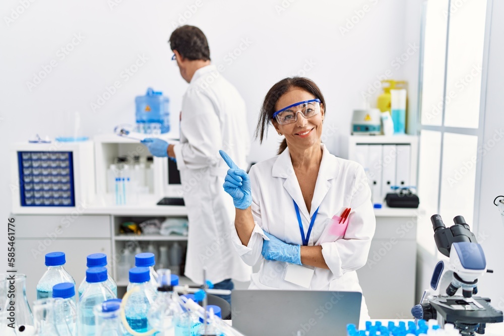 Middle age woman working at scientist laboratory smiling happy pointing with hand and finger to the side